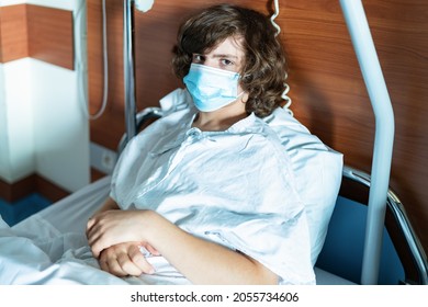 Young Teenager Boy Wearing Protection Mask Is Sitting In A Hospital Bed In A Ward. Coronavirus Infection.  Child Looking Seriously At The Camera.  Urgent Hospitalization.