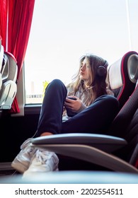 Young Teenager Girl On A Bus By A Window. Travel And Road Trip Concept. Model With Long Hair Wearing Simple Clothes And Black Earphone. Selective Focus.