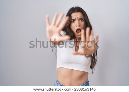 Young teenager girl standing over white background afraid and terrified with fear expression stop gesture with hands, shouting in shock. panic concept. 