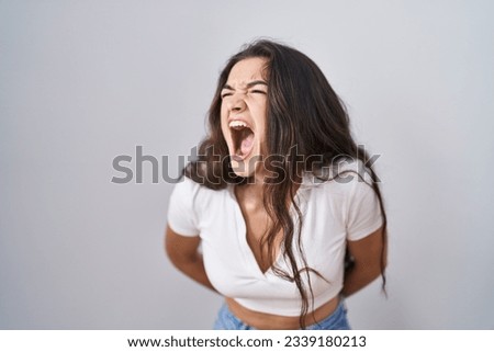 Young teenager girl standing over white background angry and mad screaming frustrated and furious, shouting with anger. rage and aggressive concept. 