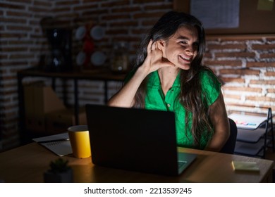 Young Teenager Girl Working At The Office At Night Smiling With Hand Over Ear Listening An Hearing To Rumor Or Gossip. Deafness Concept. 