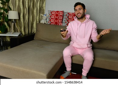 Young Television Addict Man Sitting On Sofa At Home And Watching TV Using Remote Control, Looking Excited, Pumping Out Someone Else's Comedy Comedy Movie Or Live Music At Night