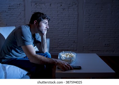 Young Television Addict Man Sitting On Home Sofa Watching TV Eating Popcorn Using Remote Control Looking Bored And Tired Zapping Foe Another Movie Sitcom Or Live Sport At Night