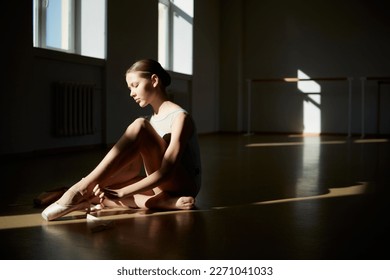 Young tender teen girl, classical ballet dancer getting ready to train in ballet school on a daytime with sunlight. Putting on pointe shoes. Ballet, dance art, education, beauty, choreography concept