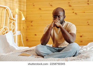 Young tense man in jeans and t-shirt keeping clenched hands by his face while sitting on bed with his legs crossed and thinking - Shutterstock ID 2258750345