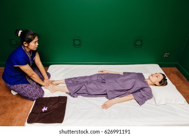 Young Thai masseur trainer girl in oriental exotic dress making and demonstrating different traditional spa ceremony procedures to european beautiful woman in pajamas in green relaxation yoga studio.