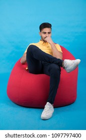 Young Thinking Man Sitting On A Red Beanbag On A Blue Background.