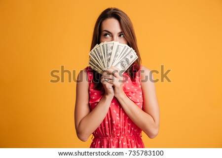 Young thinking pretty woman in red dress hiding behind bunch of money banknotes, looking upward, isolated on yellow background