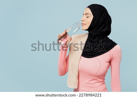 Young thirsty arabian asian muslim fitness trainer sporty woman wear pink abaya hijab towel spend time in home gym drink water isolated on plain blue background studio. Workout sport fit abs concept