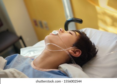 Young Thirteen Year Old Disabled Biracial Boy Lying Unconscious On Hospital Gurney Bed In Recovery Room Wearing Blue Hospital Gown, Breathing Tube Down Throat