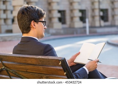 Young thoughtful businessman sitting on outdoor bench and writing in blank notepad