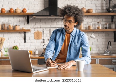 Young thoughtful casually dressed African student learning using his laptop computer while sitting at home in the cozy light colored kitchen. Distant education, e-learning, studying remotely concept - Powered by Shutterstock