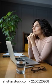 Young Thoughtful Female Employee Sitting At Office Desk With Laptop Looking Through Window, Thinking About Future Or Dreaming Of Vacation. Pensive Dreamy Businesswoman With Hands Under Chin At Work