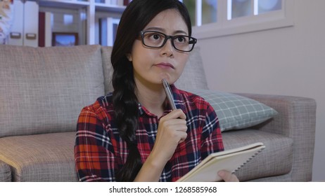 Young Thoughtful Nerd In Glasses Thinking About Test. Asian College Student Holding Pen In Chin Finding New Idea Plan For Final Semester Presentation Sitting On Floor In Apartment. Girl Stay Up Late.