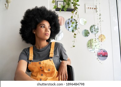 Young thoughtful pensive stylish African American gen z female hipster woman with afro hair looking through window thinking reflecting sitting lost in thoughts at home, in modern cafe alone indoors.