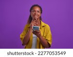 Young thoughtful positive African American woman millennial with phone in hand looks up dreamily making wish list or visualizing happy future posing on isolated lilac background. Casual girl portrait