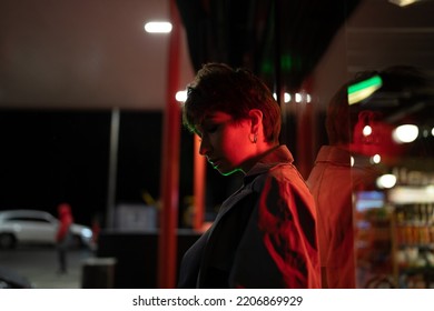 Young Thoughtful Woman Against Glass Wall Standing On Street With Red Neon Light At Night. Pensive German Girl With Short Hair Looks Ahead. Sad And Lonely Female In Big City. Sadness And Melancholy