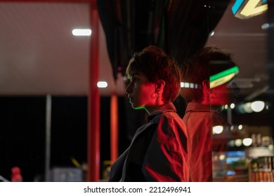 Young Thoughtful Woman Against Glass Wall Standing On Street With Red Neon Light At Night. Pensive German Girl With Short Hair Looks Ahead. Sad And Lonely Female In Big City. Sadness And Melancholy