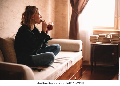 Young Thoughtful Woman Drinking Tea And Eating Chocolate While Looking Through Window Sitting On Sofa In Living Room At Home