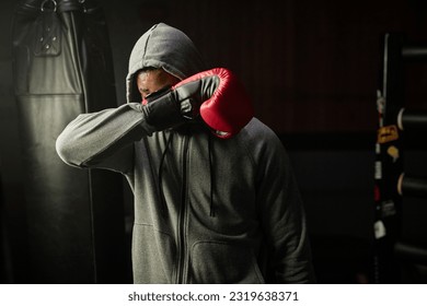 Young tired African American boxer in grey hoodie hiding his face behind red boxing glove while holding it in front of himself in sports club - Powered by Shutterstock