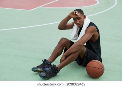 Young tired african sportsman sitting on sports court with basketball ball and wiping forehead after training. Black man with towel wear sportswear and sneakers. Urban basketball player. Daytime