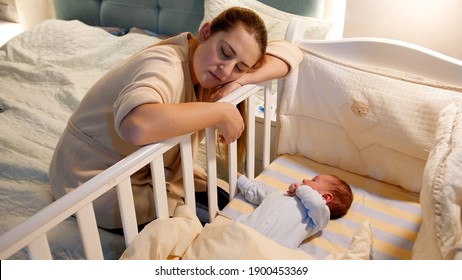 Young tired and exhausted mother fallen asleep while rocking crib of her newborn baby at night. Concept of sleepless nights and parent depression after childbirth.