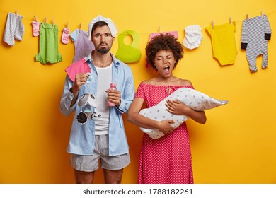 Young tired exhausted parents spend sleepless night caring about infant, nursing baby. Sleepy mother yawns and holds child on hands wrapped in blanket, displeased father holds bottle and mobile toy