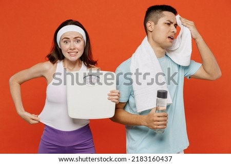 Young tired fitness trainer instructor sporty two man woman in headband t-shirt hold scales water bottle spend weekend in home gym isolated on plain orange background Workout sport lifestyle concept
