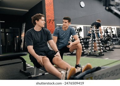 Young tired man in conversation with personal trainer in gym after workout training on rowing machine. Sweaty male taking break after workout exercise. Exhausted people talking to each others. - Powered by Shutterstock