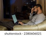 Young tired man using laptop in penthouse or luxury hotel room. Businessman in skyscraper apartment against night city background working late at nigh.