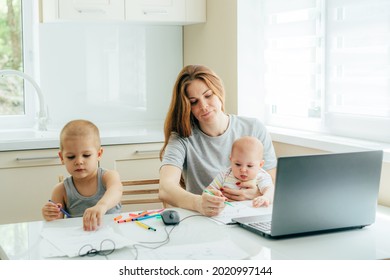 Young Tired Mom Works On A Laptop And Babysits Children In The Kitchen At The Table.