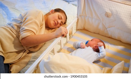 Young Tired Mother Fallen Asleep While Rocking Her Baby Son In Crib At Late Night. Motherhood And Sleepless Nights