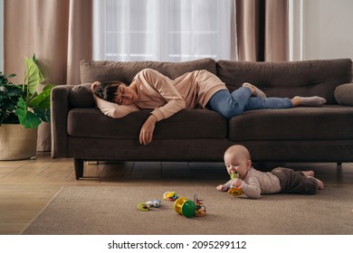 Young tired mother suffering from lack of sleep, sleeping on sofa while her little infant baby is playing on floor. Exhausted mom experiencing postnatal depression, does not want to play with her son