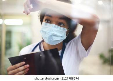 Young Tired Overworked African American Medical Worker Wearing Protective Face Mask With X-ray Scan In Hands Looking Out Window With Frustrated Expression. Healthcare Workers During COVID-19 Pandemic