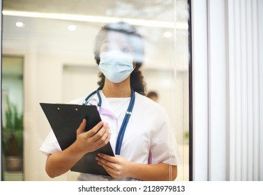 Young Tired Overworked African American Medical Worker Wearing Protective Face Mask With X-ray Scan In Hands Looking Out Window With Frustrated Expression. Healthcare Workers During COVID-19 Pandemic