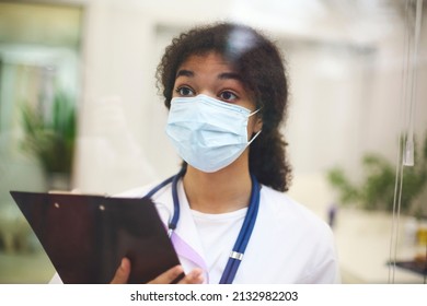 Young Tired Overworked African American Medical Worker Wearing Protective Face Mask With X-ray Scan In Hands Looking Out Window With Frustrated Expression. Healthcare Workers During COVID-19 Pandemic