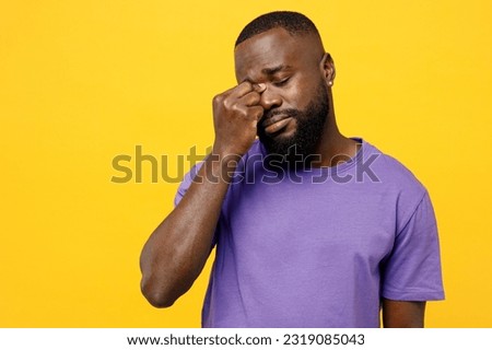 Young tired sad ill man of African American ethnicity he wear casual clothes purple t-shirt keeping eyes closed rub put hand on nose worry isolated on plain yellow background studio. Lifestyle concept