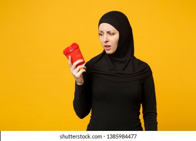 Young Tired Sad Upset Arabian Muslim Woman In Hijab Black Clothes Hold Paper Cup Of Coffee Isolated On Yellow Wall Background, Studio Portrait. People Religious Lifestyle Concept. Mock Up Copy Space