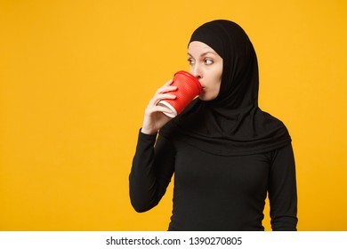 Young Tired Sad Upset Arabian Muslim Woman In Hijab Black Clothes Hold Paper Cup Of Coffee Isolated On Yellow Wall Background, Studio Portrait. People Religious Lifestyle Concept. Mock Up Copy Space