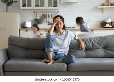 Young tired single mother suffers from headache closed eyes touch forehead sitting on couch while her daughter and son running around her and shouting, female babysitter feels exhausted by noisy kids - Shutterstock ID 1606546576