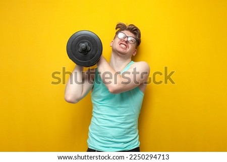 young tired thin guy nerd lifts heavy dumbbells and cries on a yellow background, a confused student in glasses is training