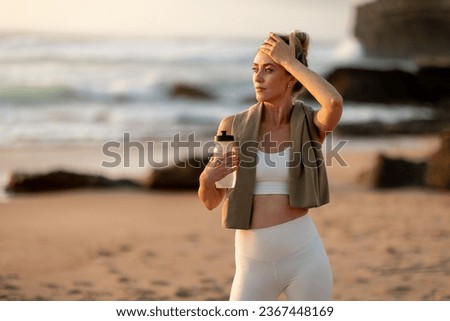 Young tired woman athlete in sportswear holding bottle of water, resting after workout on sea beach, free space. Break, active lifestyle, health care, sport and aqua balance