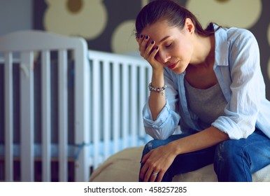 Young tired woman sitting on the bed near children's cot.