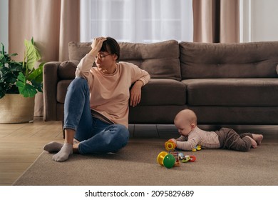 Young tired woman suffering from postnatal depression, holding her head with hand, sitting on floor in living room near her toddler baby playing with toys. New mom tired from motherhood difficulties