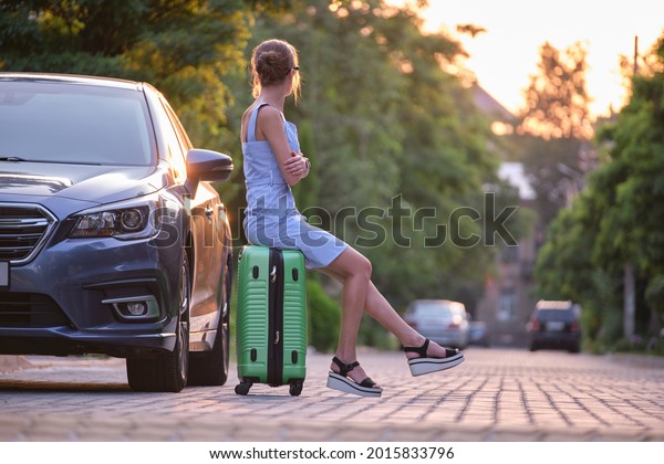 Young tired woman with\
suitcase sitting beside car waiting for someone. Travelling and\
vacations concept.