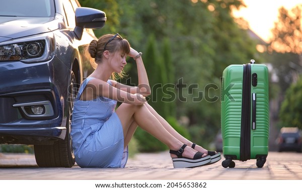Young tired woman with\
suitcase sitting beside car waiting for someone. Travelling and\
vacations concept.