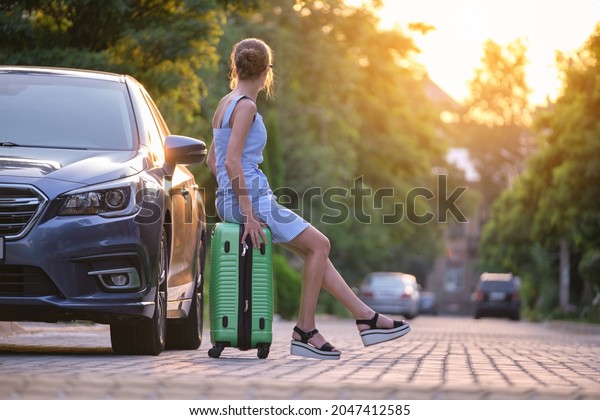 Young tired woman with\
suitcase sitting near her car waiting for someone. Travel and\
vacations concept.