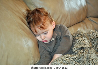 Young Toddler Girl With Long Eyelashes, Light Red Hair, Red Pout Lips, Wearing Brown Long Sleeved Shirt, Sleeping On A Tan Leather Couch While Covered In A Brown Knit Blanket, Arms Stretched Out.