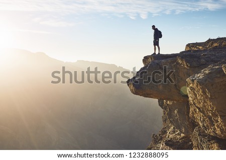 Young tourist with backpack standing on the edge of cliff at sunrise. Jebel Akhdar, Grand Canyon of Oman.  
