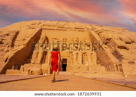 A young tourist girl in red dress walking towards the Abu Simbel Temple in southern Egypt in Nubia next to Lake Nasser at sunset. Temple of Pharaoh Ramses II